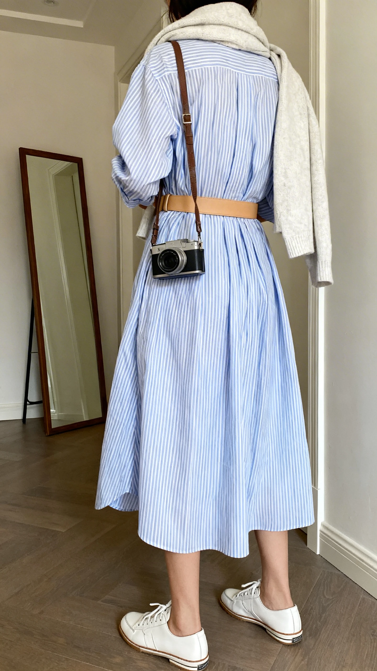 iPhone back view of a woman wearing a breezy blue-and-white striped shirt dress cinched with a tan belt, white leather sneakers, crossbody camera bag, and a lightweight sweater tossed over shoulders, face not visible, hallway full-length mirror, natural daylight, iPhone photo quality