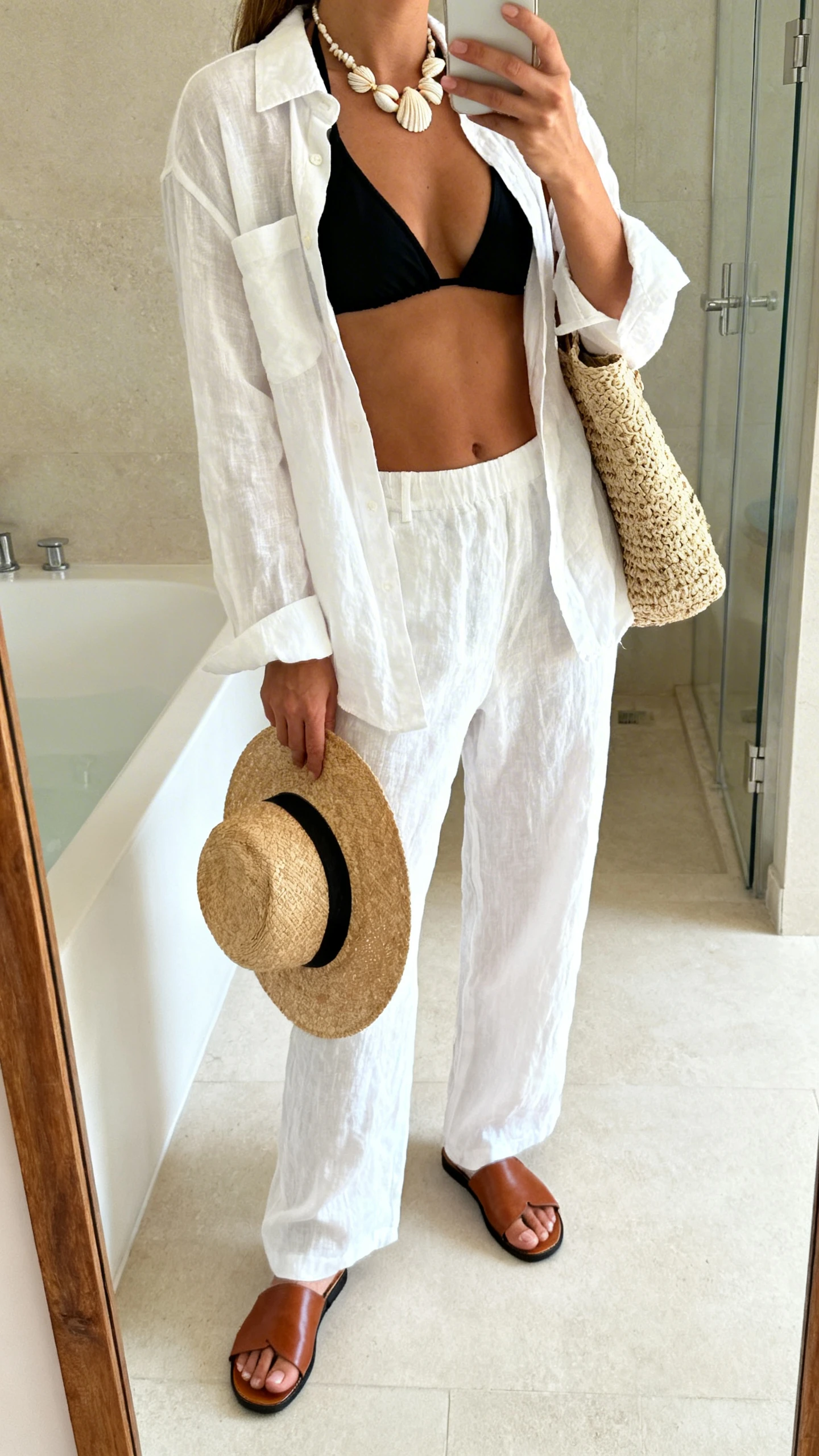 iPhone mirror selfie of a woman wearing a chic black bikini top under an open white linen shirt with matching linen pants, leather slide sandals, a straw hat held in hand, shell necklace, and woven tote, face not visible, side angle, bathroom mirror, bright natural light, iPhone photo quality